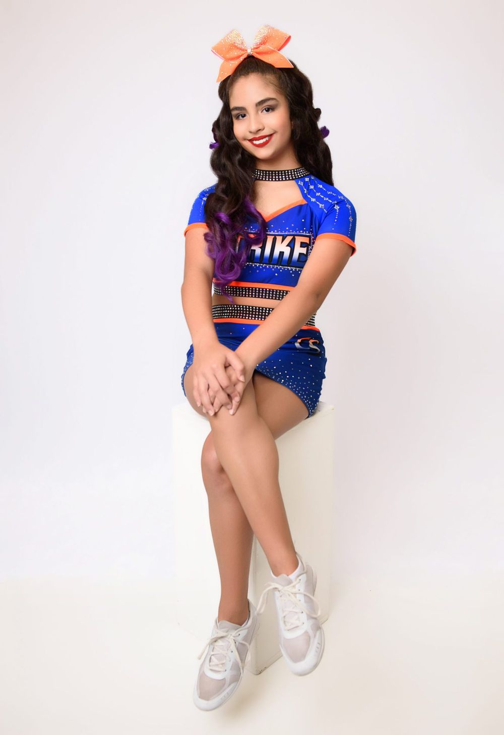 Cheerleader in blue uniform with orange bow poses in studio setting.