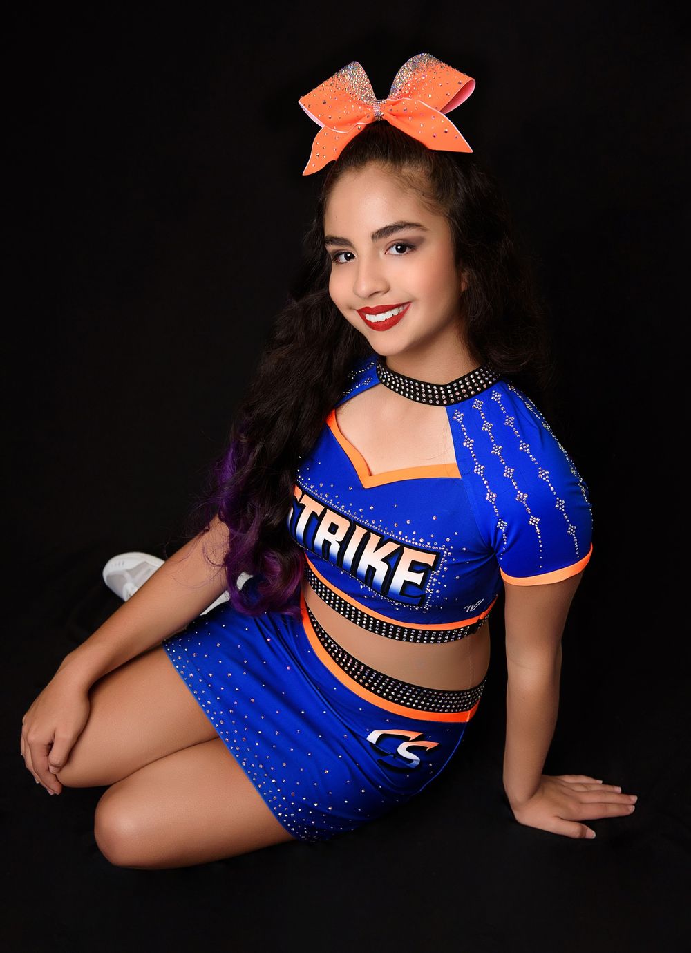 A cheerleader in sparkly blue uniform strikes a pose while wearing an orange bow.