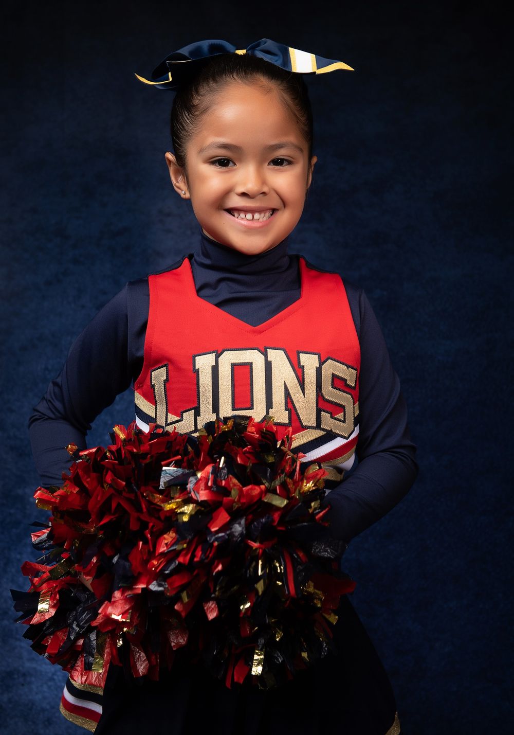A cheerleader in a Lions uniform smiles while holding red pom poms against a blue backdrop.