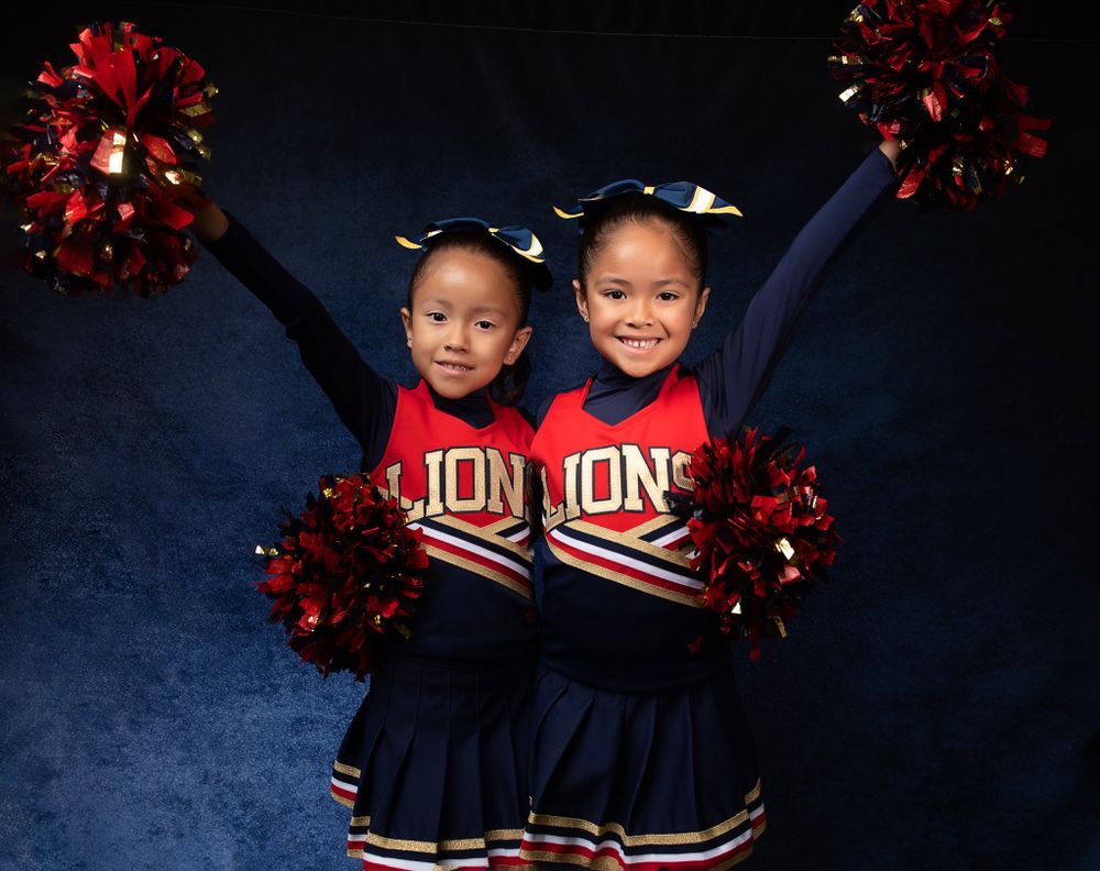 Two cheerleaders in Lions uniforms raise their pom poms high against a blue background.