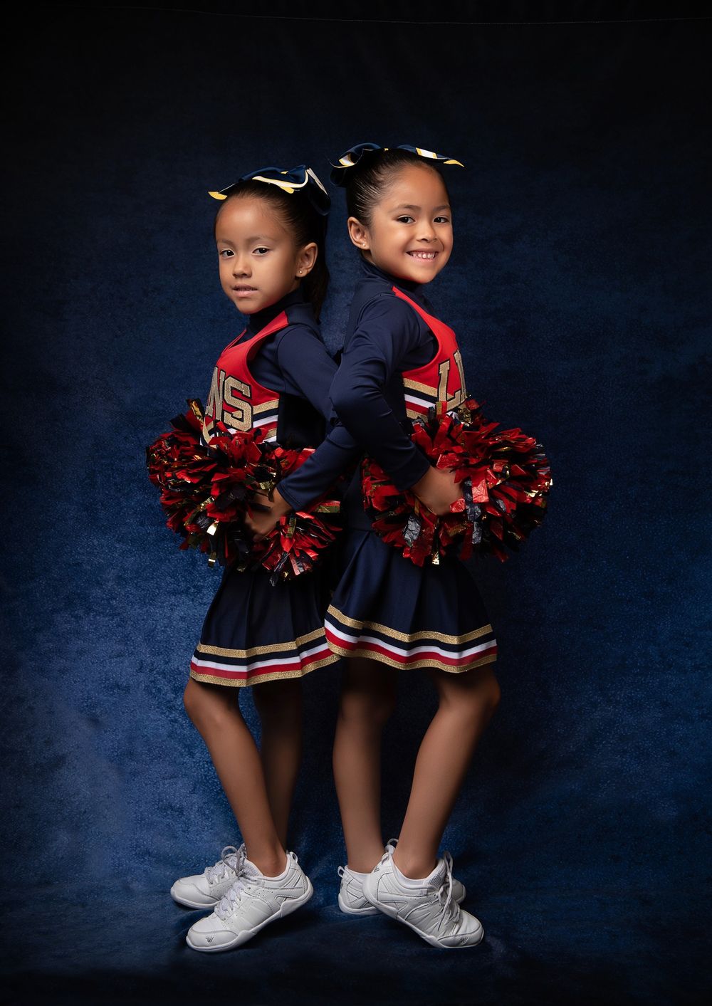 Two young cheerleaders in navy uniforms with red pom poms pose back-to-back against a blue backdrop.