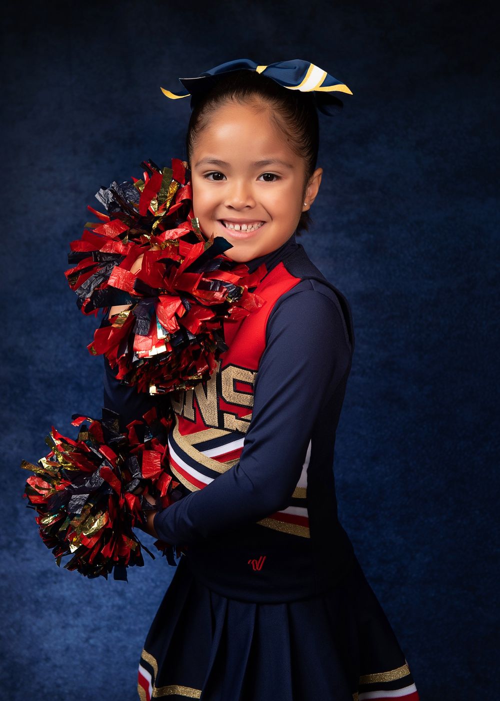 A cheerleader in a navy and red uniform holds red pom poms against a dark blue background.