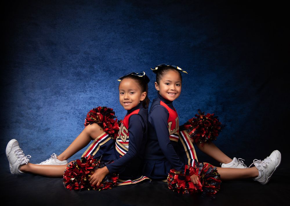 Two cheerleaders in matching uniforms sitting on the floor with red pom poms against a dark backdrop.