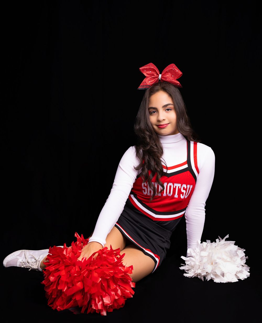 Cheerleader in red and black uniform poses seated with pom poms against black backdrop.