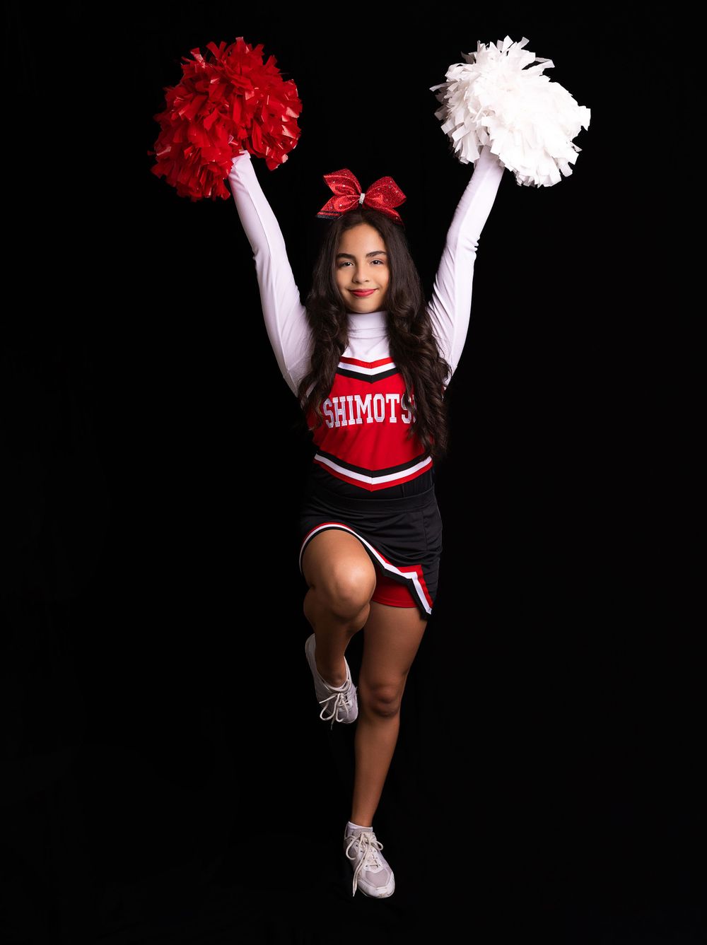 Cheerleader in red and black uniform jumps with red and white pom poms against dark background.