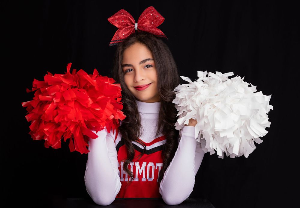 A cheerleader in uniform poses with red and white pom poms against black background.