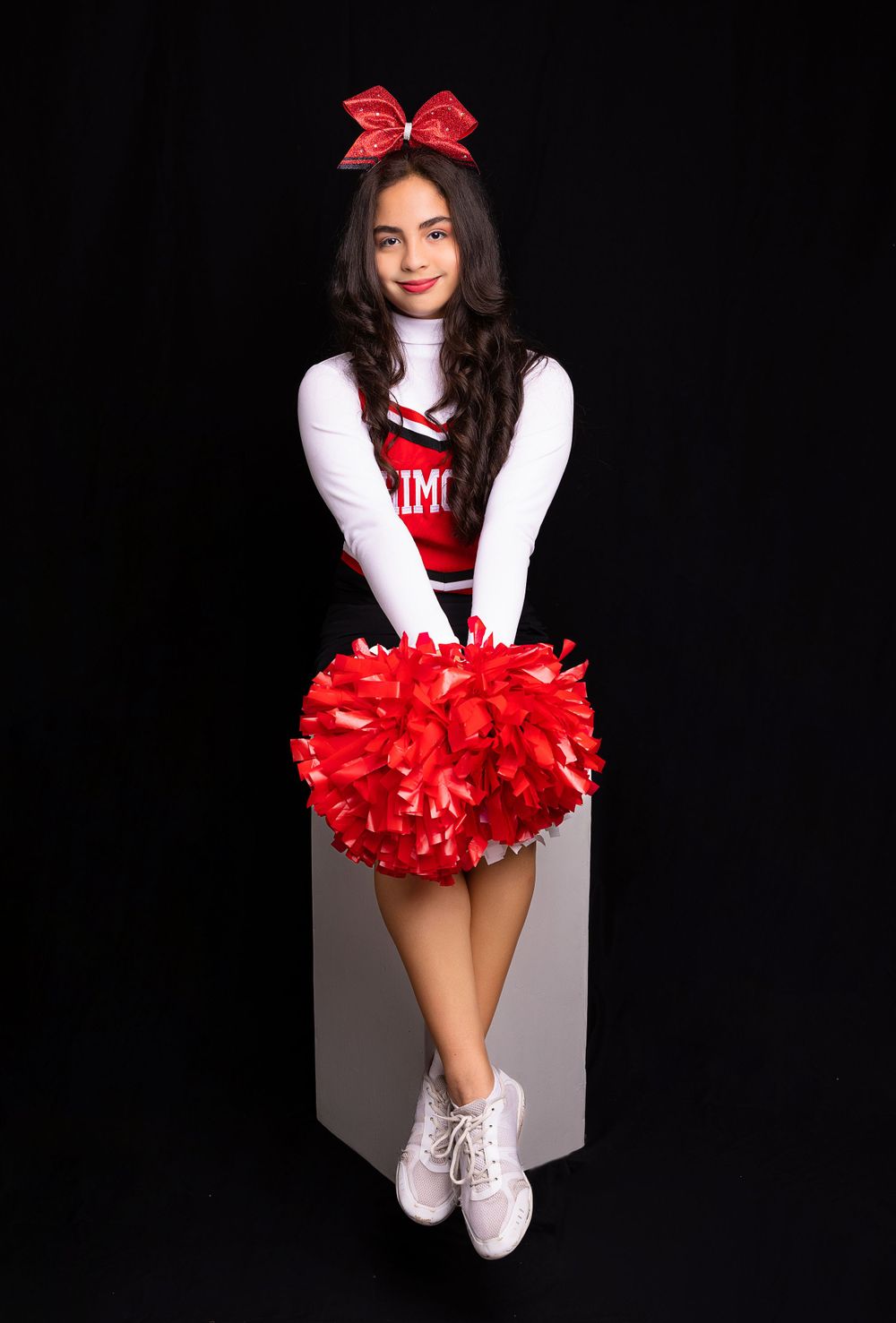 Cheerleader in black and red uniform sits with red pom pom on gray block.