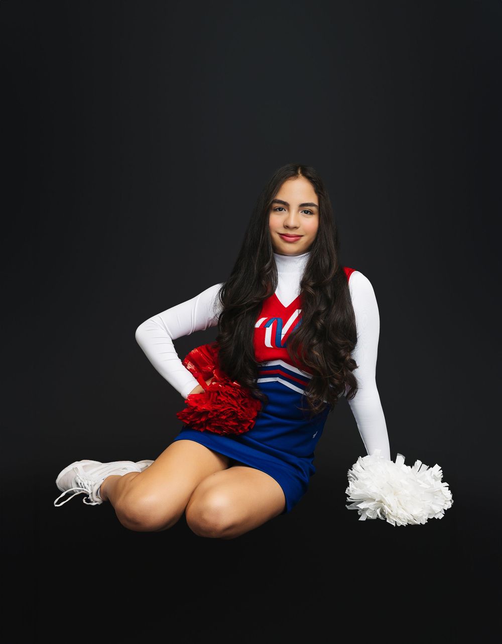 Cheerleader in blue and red uniform sits with pom poms against dark background.