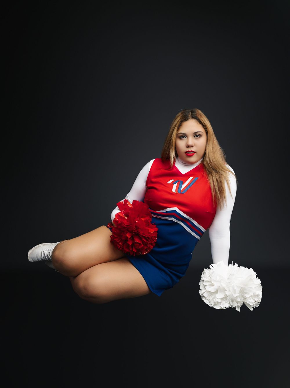 Athletic cheerleader in red and blue uniform poses with pom poms against dark background.