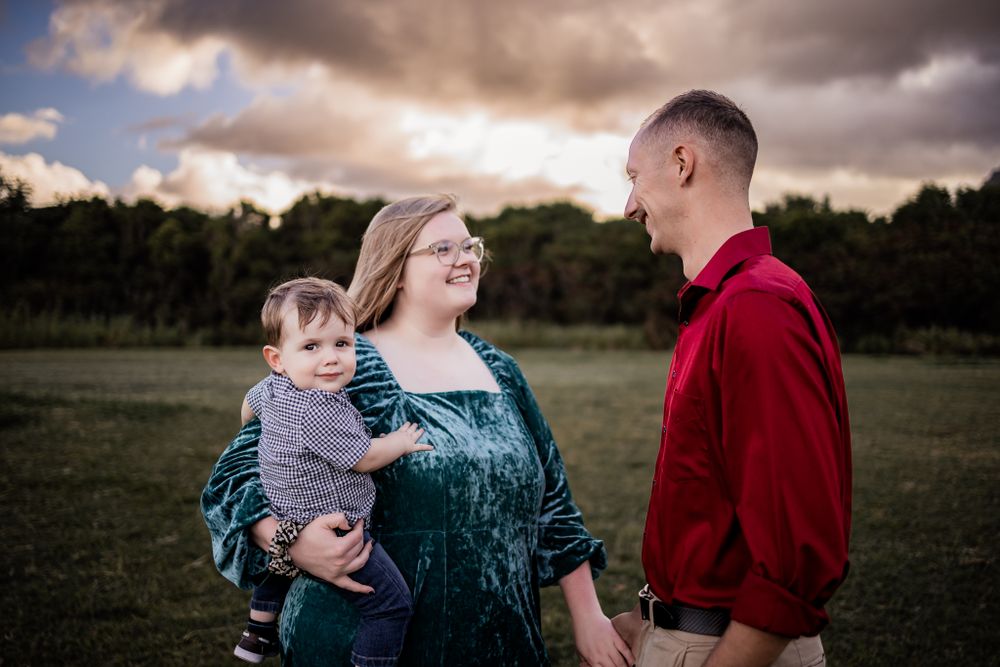 A family stands together in a field beneath dramatic cloudy skies at sunset.