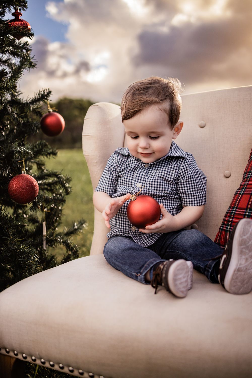 A toddler in a checkered shirt plays with red Christmas ornaments while sitting in a beige chair.