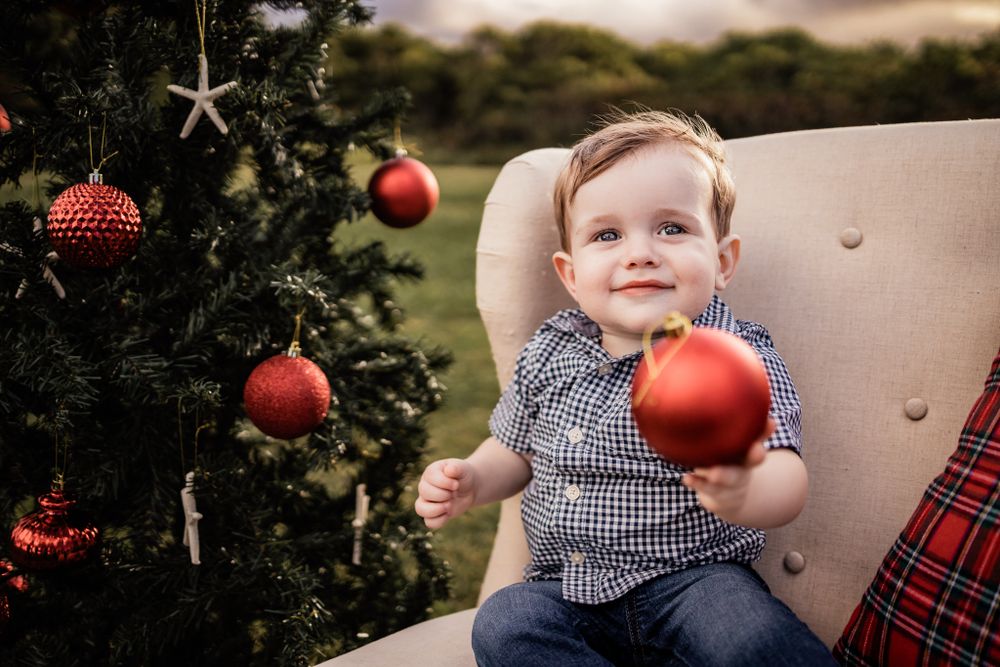 A child smiles while holding a red Christmas ornament next to a decorated tree.