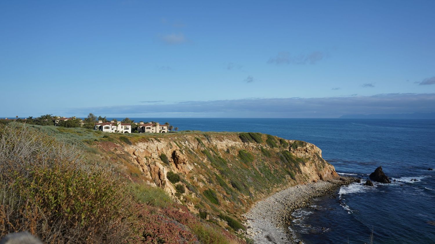 Scenic coastal cliffside with houses perched on rocky bluffs overlooking calm blue Pacific Ocean.