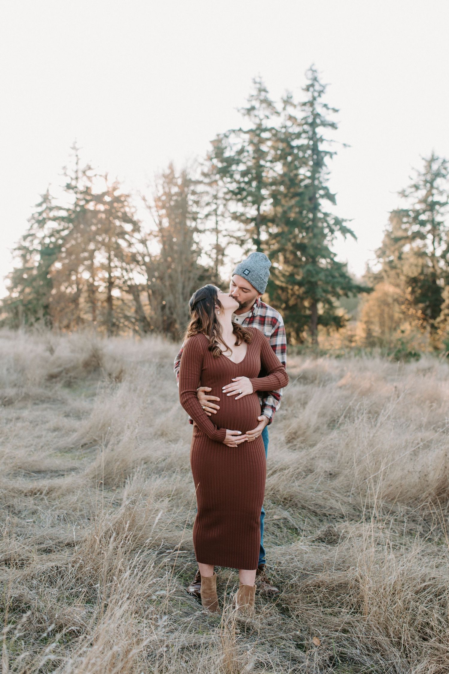 Couple sharing intimate moment in field of tall grass with evergreen trees in background.