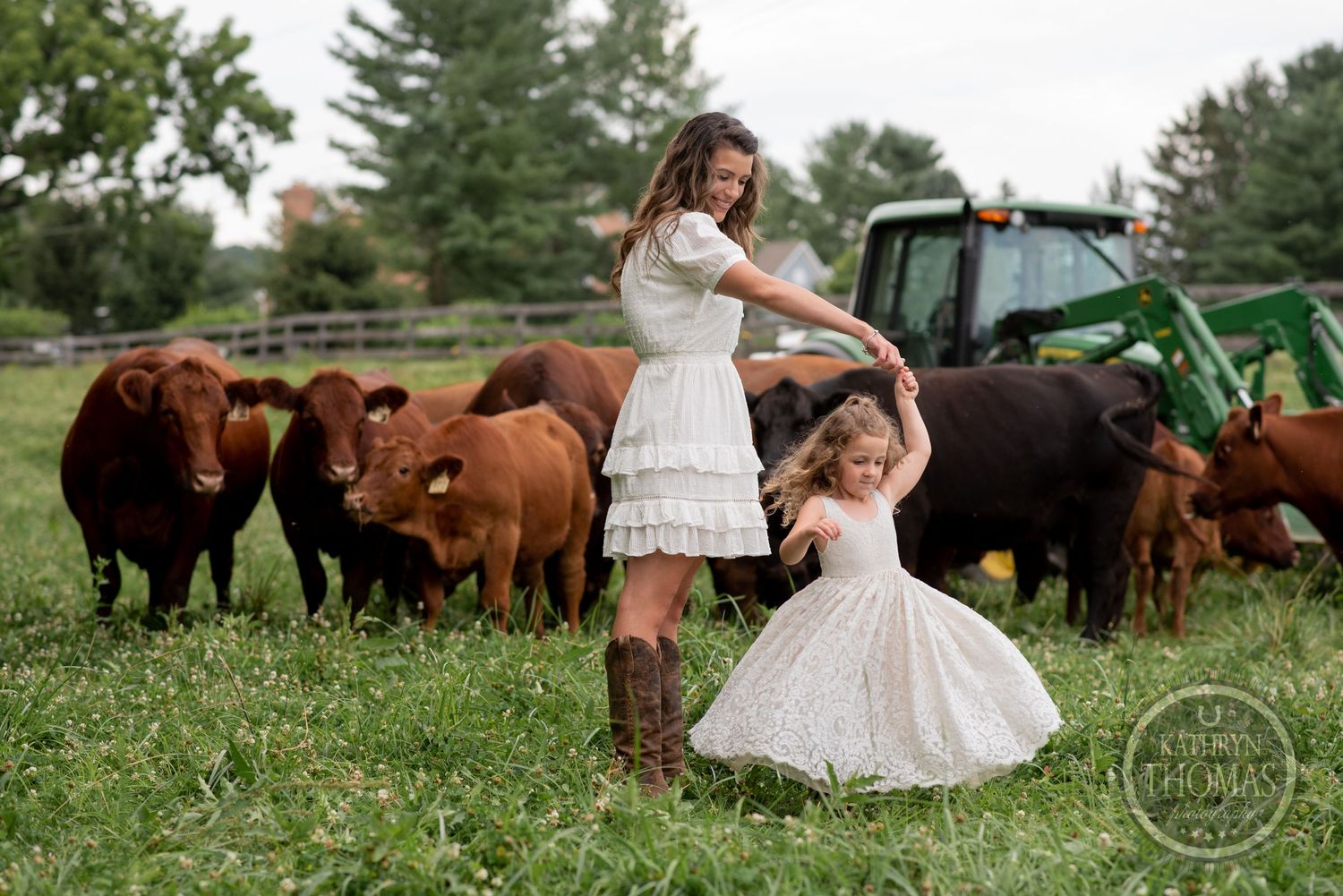 Two people in white dresses dance playfully in a field with cattle and a green tractor behind them.
