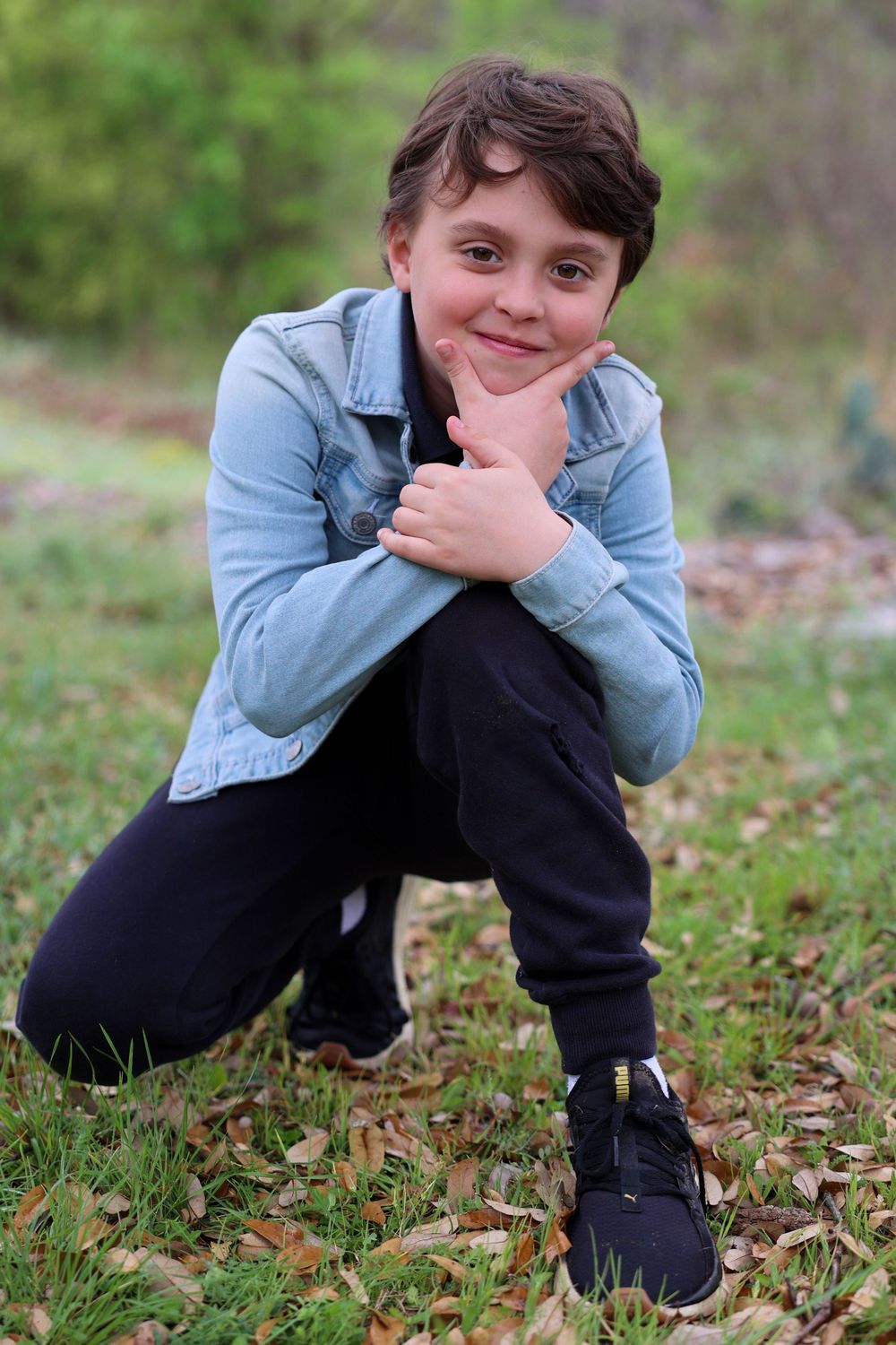 Young person in denim jacket crouches while posing playfully outdoors.