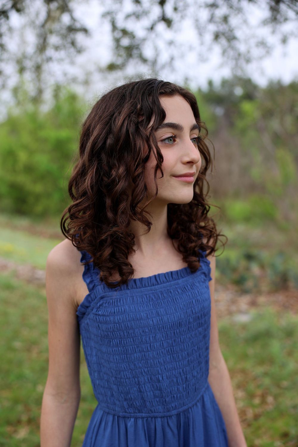 Young woman in blue dress with curly dark hair poses in outdoor garden setting.