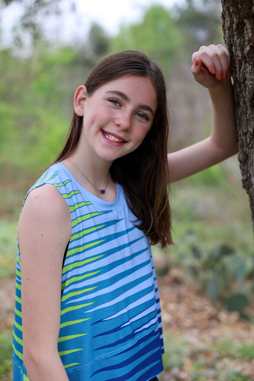 Portrait of a young person in blue striped tank top leaning against tree trunk outdoors.