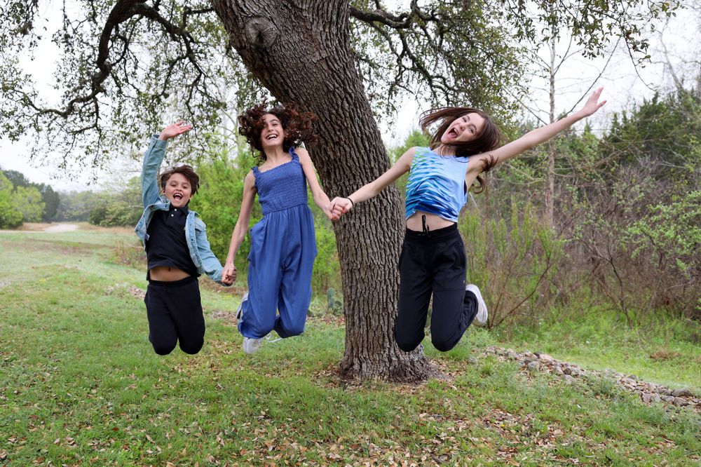 Three people jumping in unison next to a large oak tree on a sunny day.