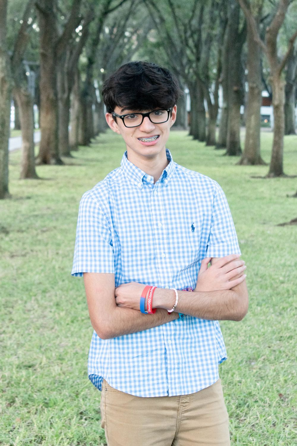 A young person in a blue gingham shirt and glasses stands in a tree-lined path.