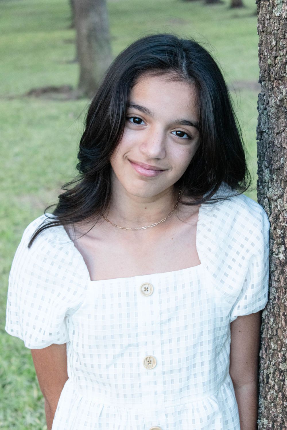 A young woman in a white textured dress poses against a tree in a park setting.