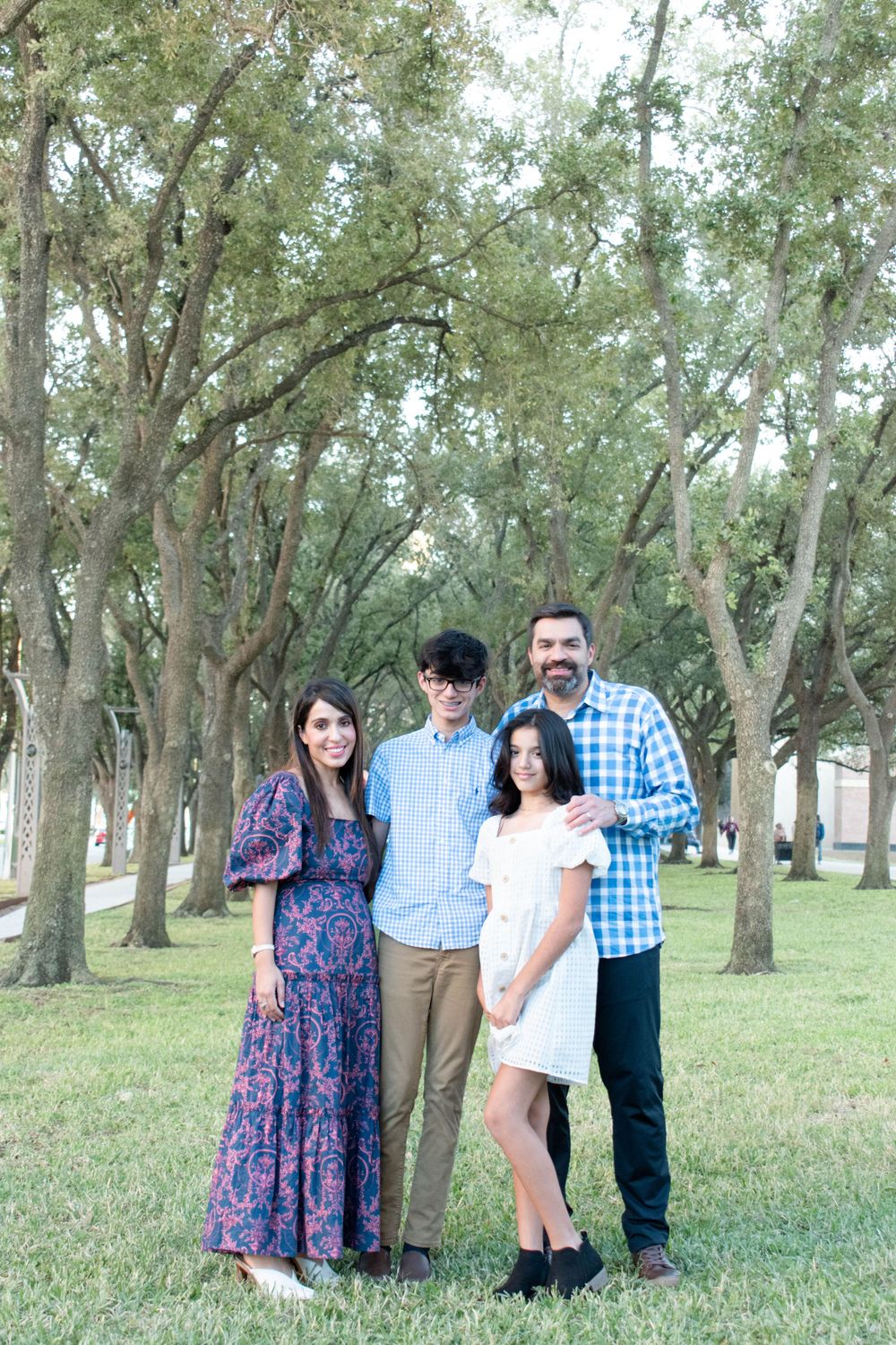 A family of four poses together outdoors in a tree-lined park with coordinated blue outfits.