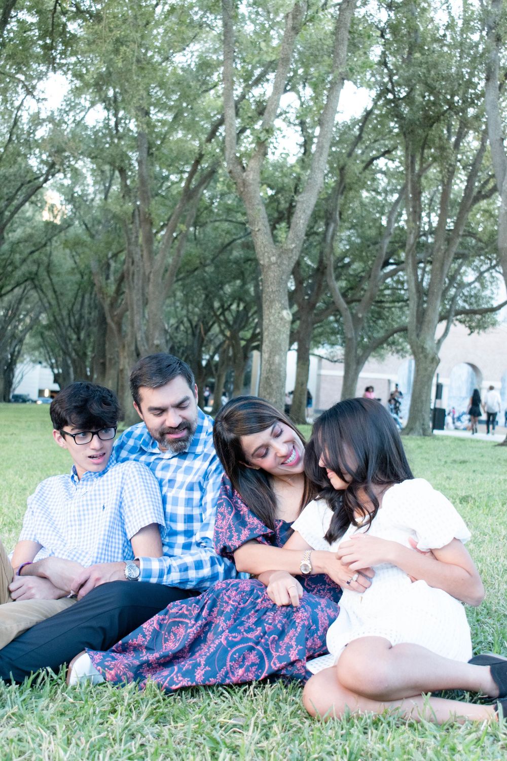 A family shares a candid laughing moment while sitting together on grass under shady trees.
