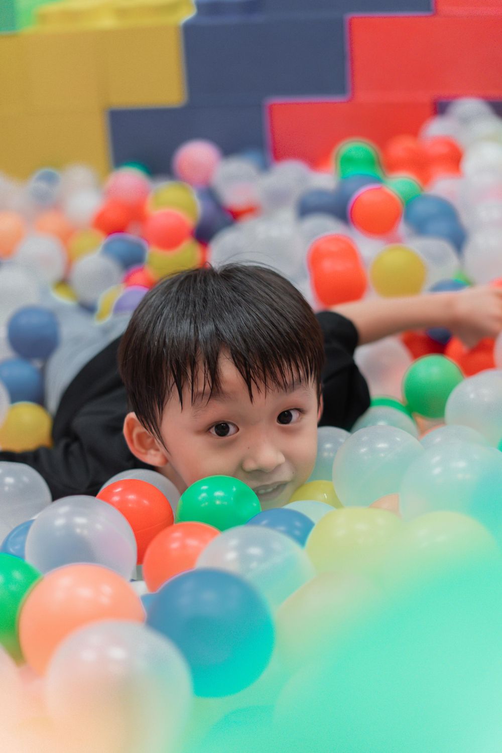 Child peeking through colorful balls in a ball pit with building blocks in the background.