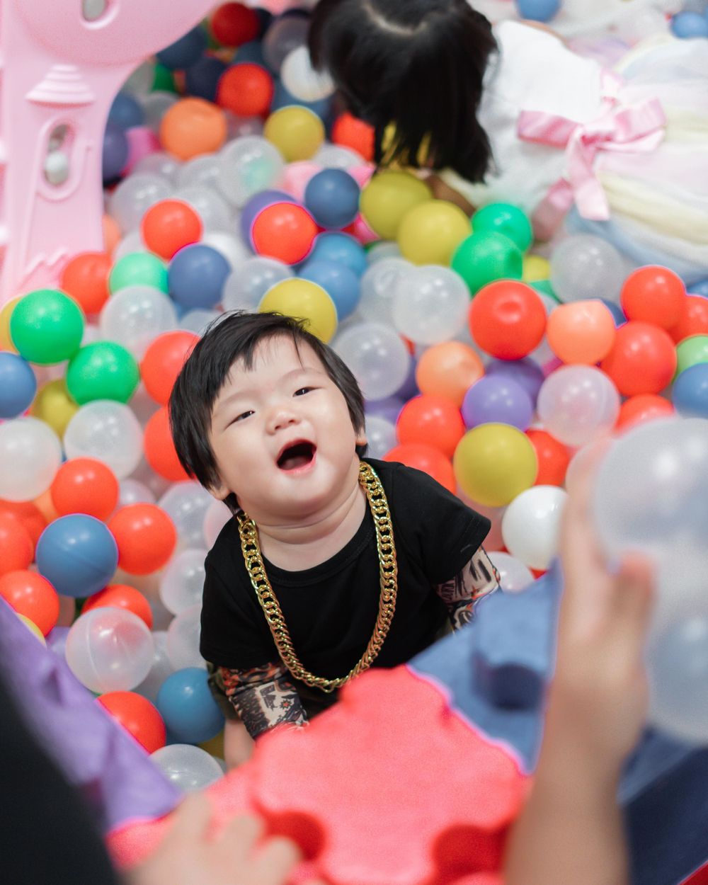 Child playing joyfully in a colorful ball pit wearing a gold chain necklace.