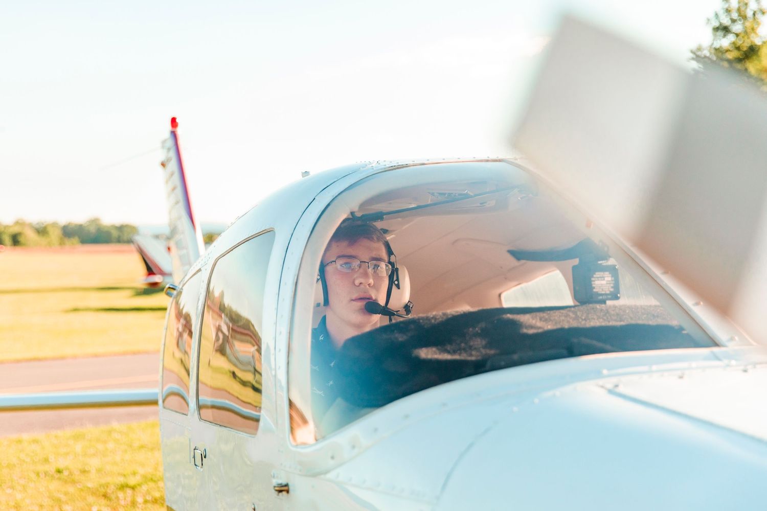 Young pilot wearing headset sits in cockpit of small white aircraft on sunny airfield.