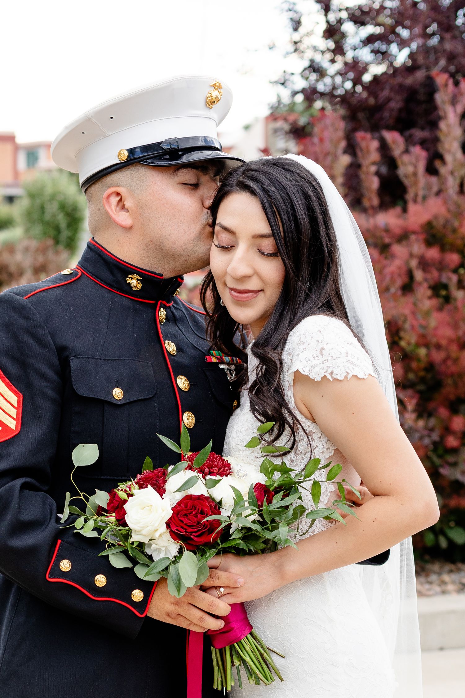 Marine in dress uniform embraces bride holding red and white bouquet against autumn foliage backdrop.