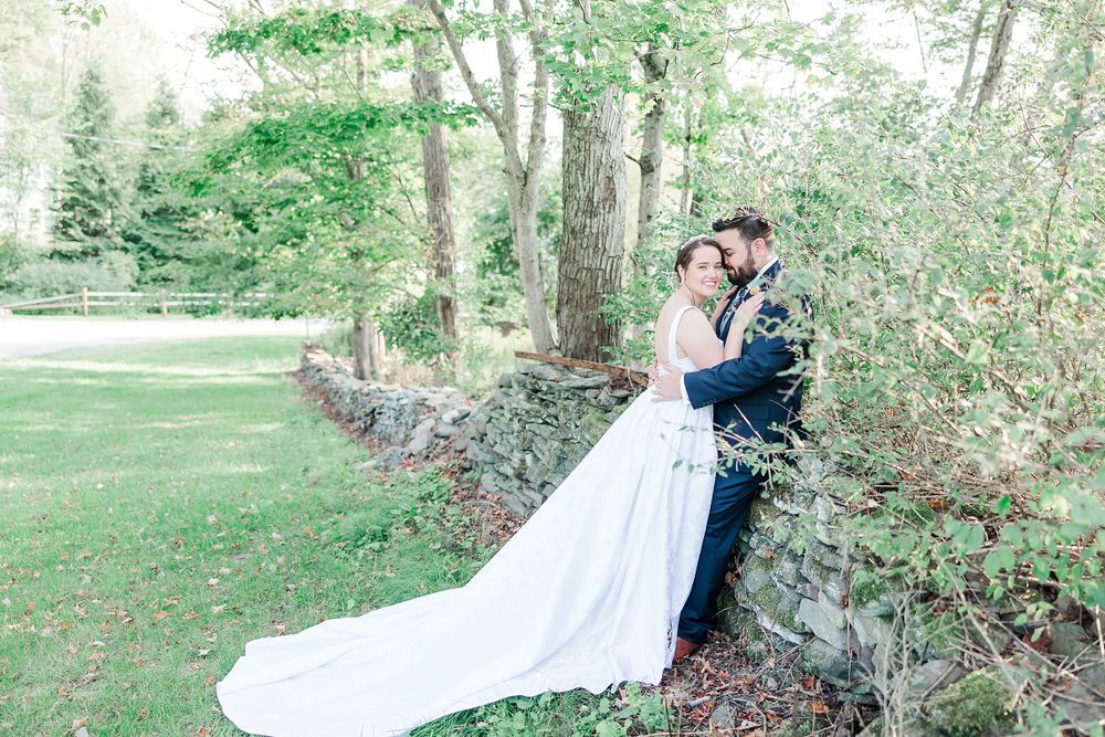 Wedding couple embraces near rustic stone wall surrounded by green forest scenery.