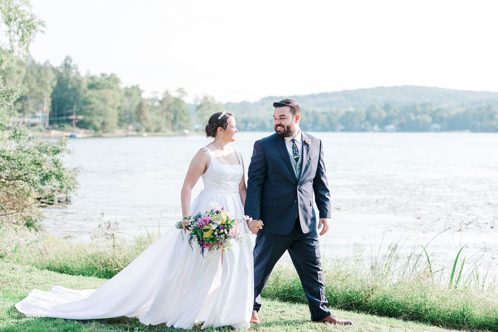 Couple in wedding attire poses by scenic lake with mountains in background.