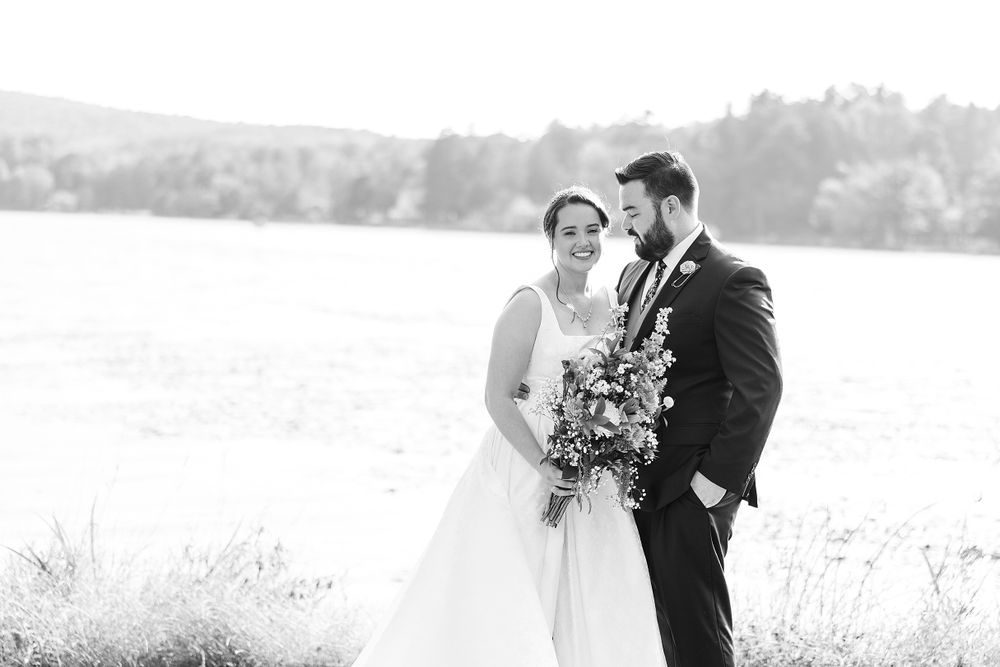 Black and white wedding portrait of couple with floral bouquet against scenic lakeside backdrop.