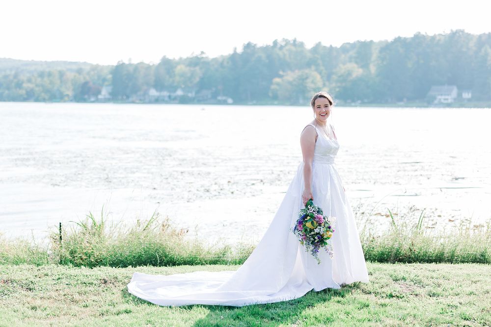 Bride in flowing white gown holds bouquet while posing on lakeside lawn.