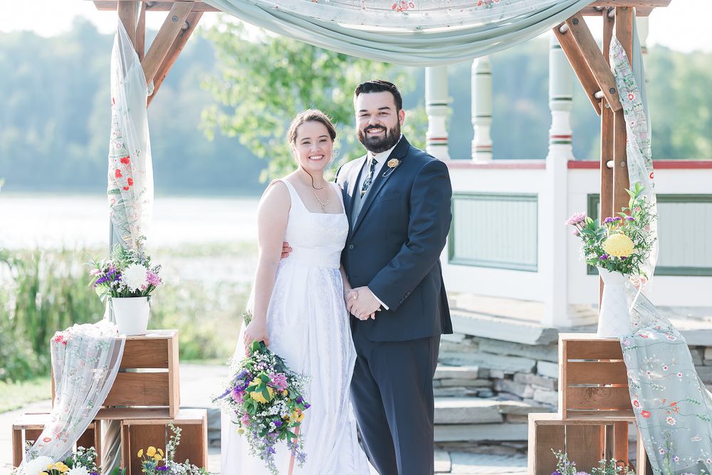 Newlyweds pose under decorative gazebo with lake and floral arrangements in background.
