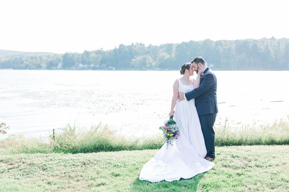 Lakeside wedding portrait with couple embracing on grassy shore with water views.