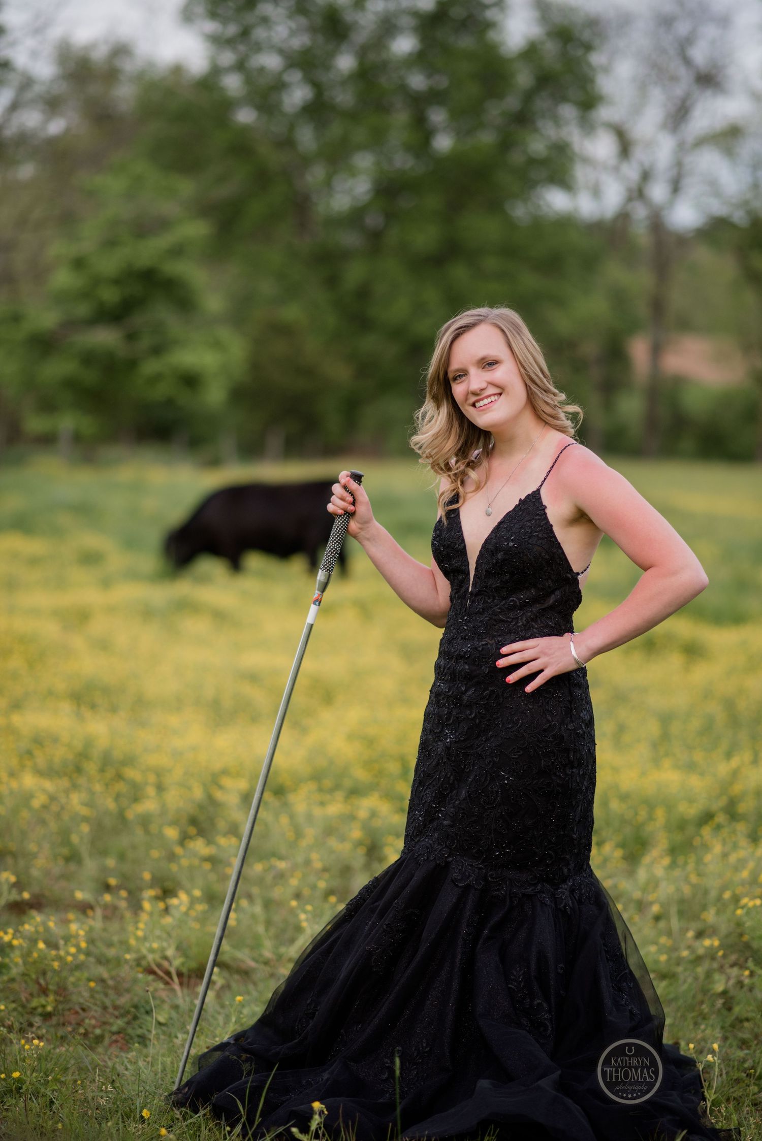 A formal portrait in a meadow features a black mermaid gown and holding a silver staff against yellow wildflowers.