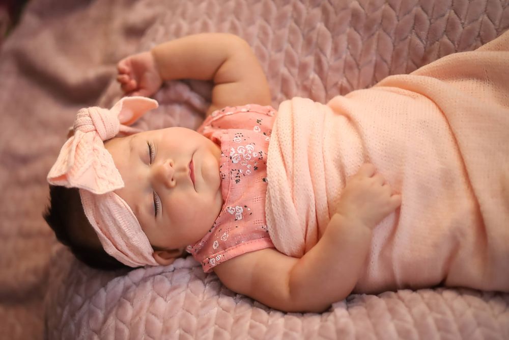 Baby in pink outfit and bow lying on textured fabric.