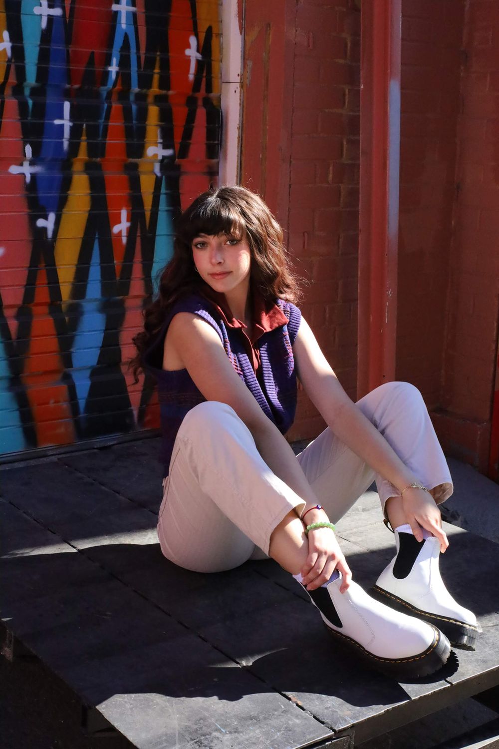 Fashion shoot against colorful street art wall featuring casual urban style with white boots and purple vest.