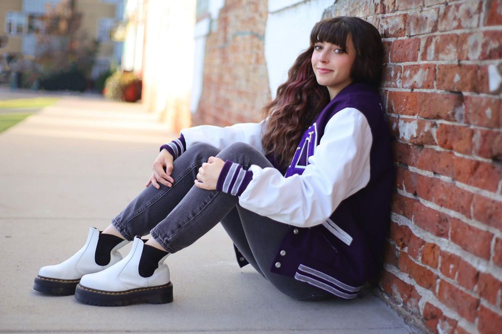 Casual urban portrait against brick wall wearing varsity jacket and white boots.