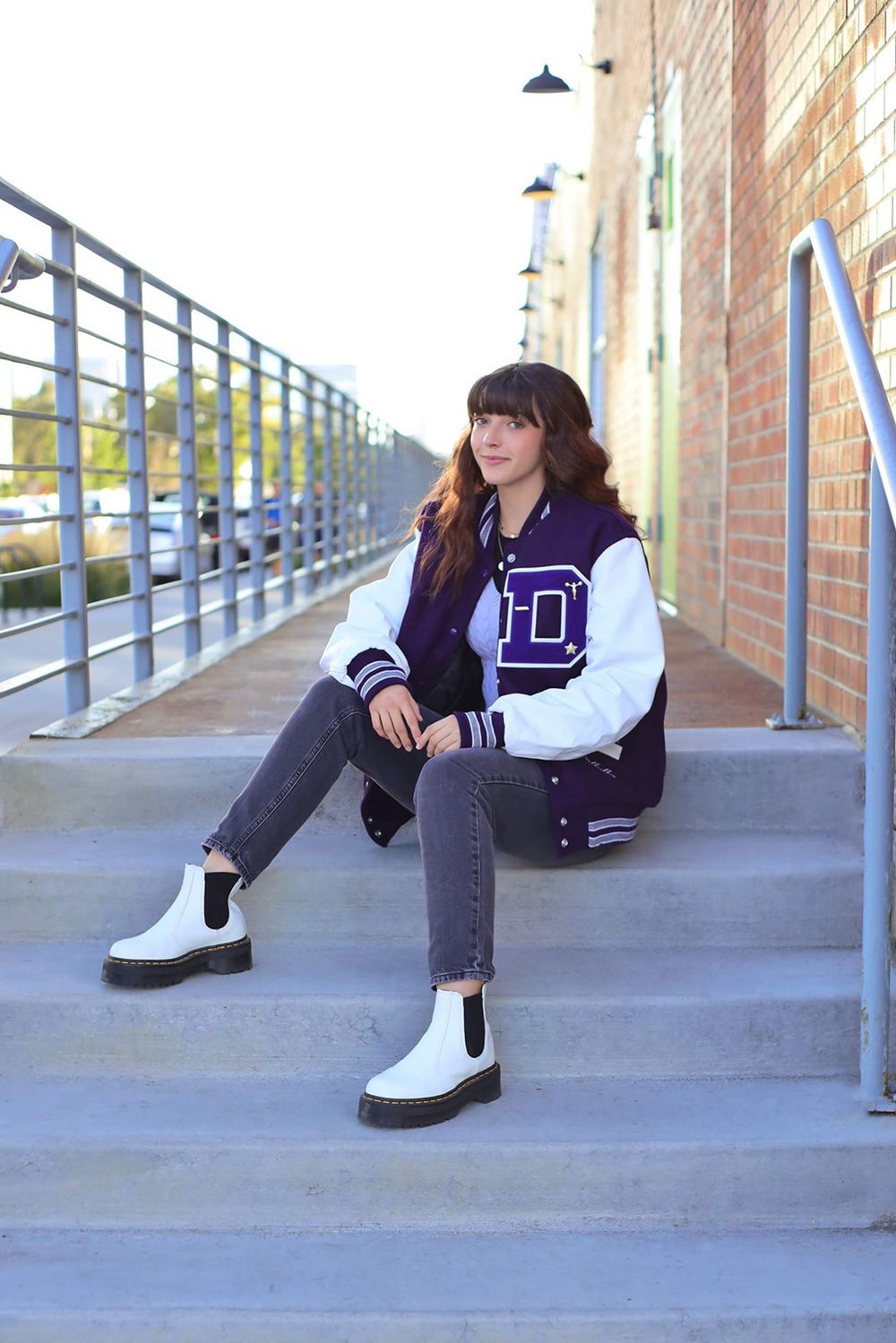 Street style portrait on concrete steps wearing varsity jacket and white boots.