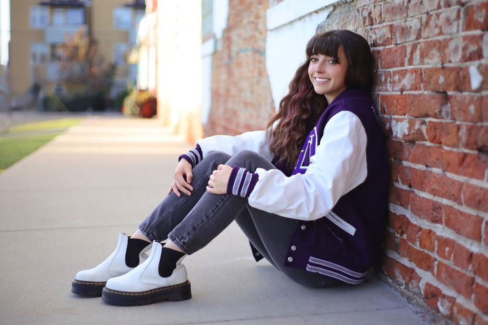 Relaxed outdoor portrait wearing purple and white varsity jacket against brick wall.