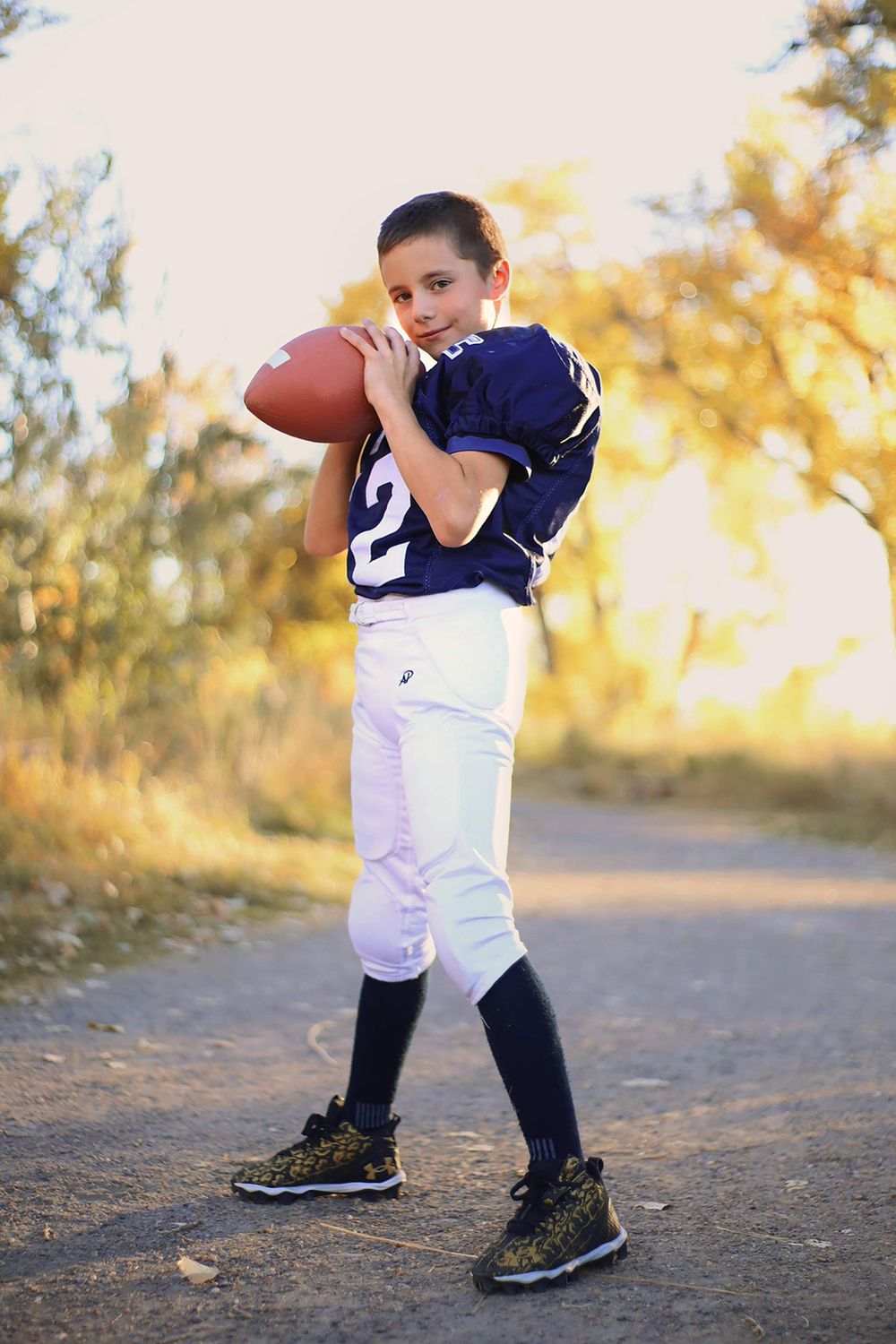 Youth football player in navy jersey practices throwing technique on outdoor path with autumn foliage background.