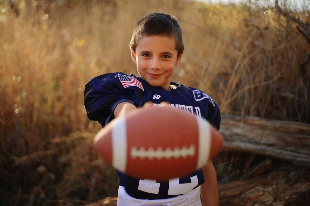 Youth football player holding ball in navy uniform poses at sunset.