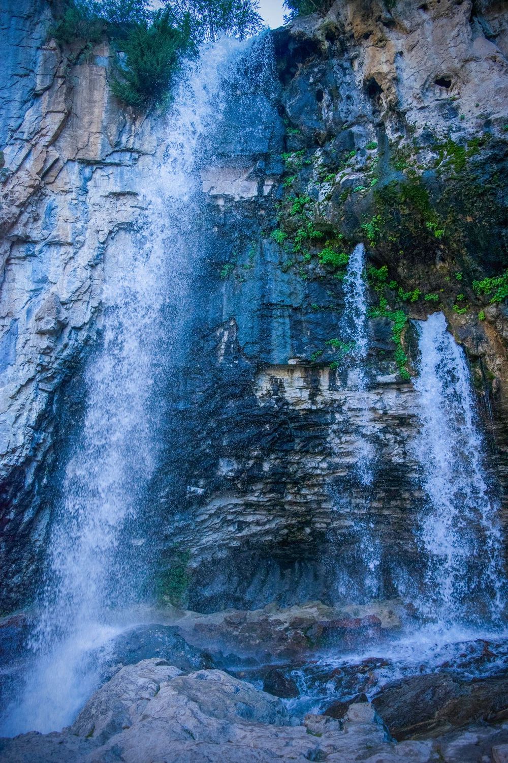 Tall waterfall cascades down layered limestone cliff face surrounded by green vegetation.
