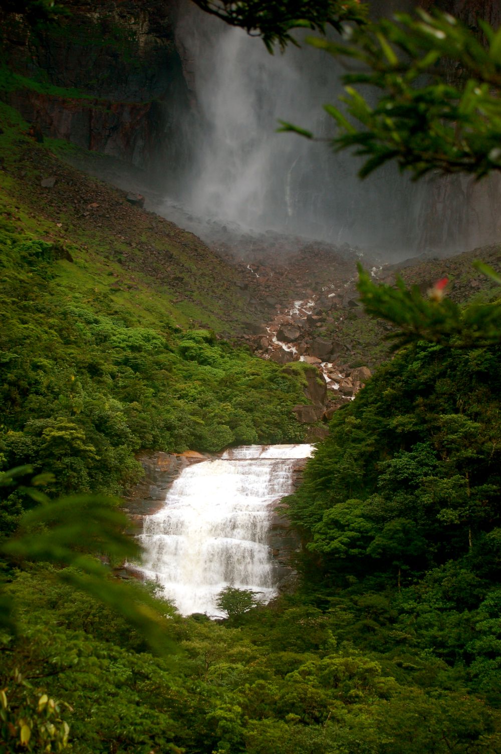 A majestic two-tiered waterfall cascades down a lush green mountainside surrounded by tropical vegetation.