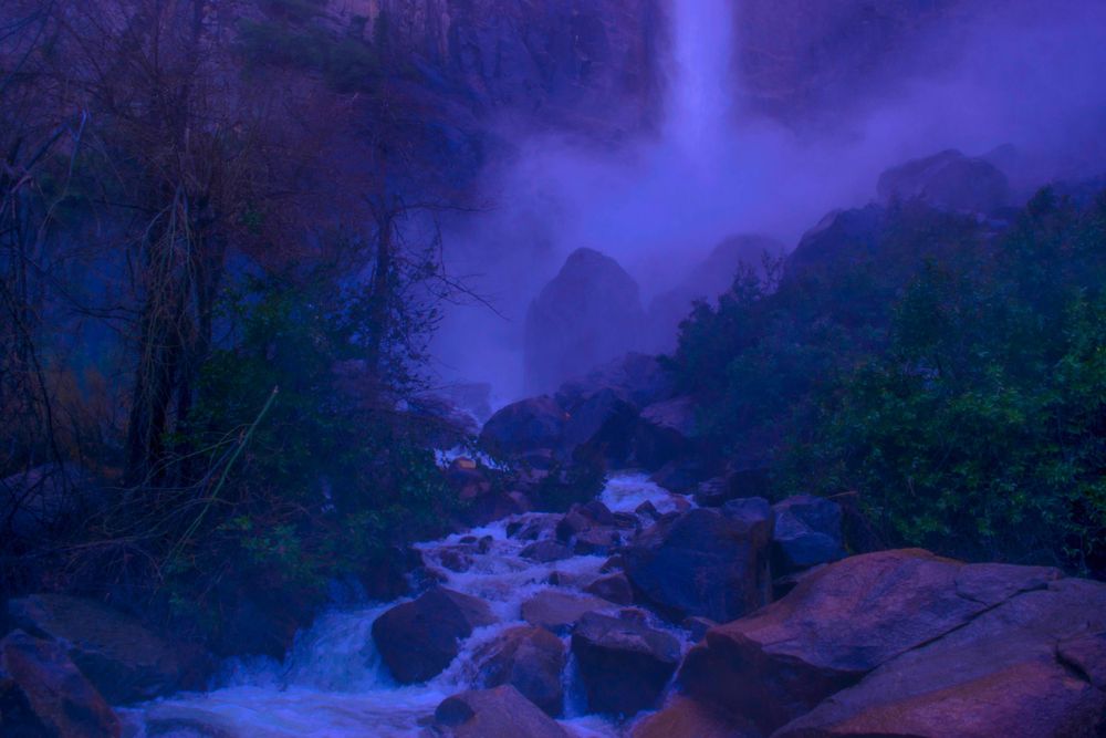 Mystical blue-tinted waterfall scene with rocky stream and misty atmosphere.