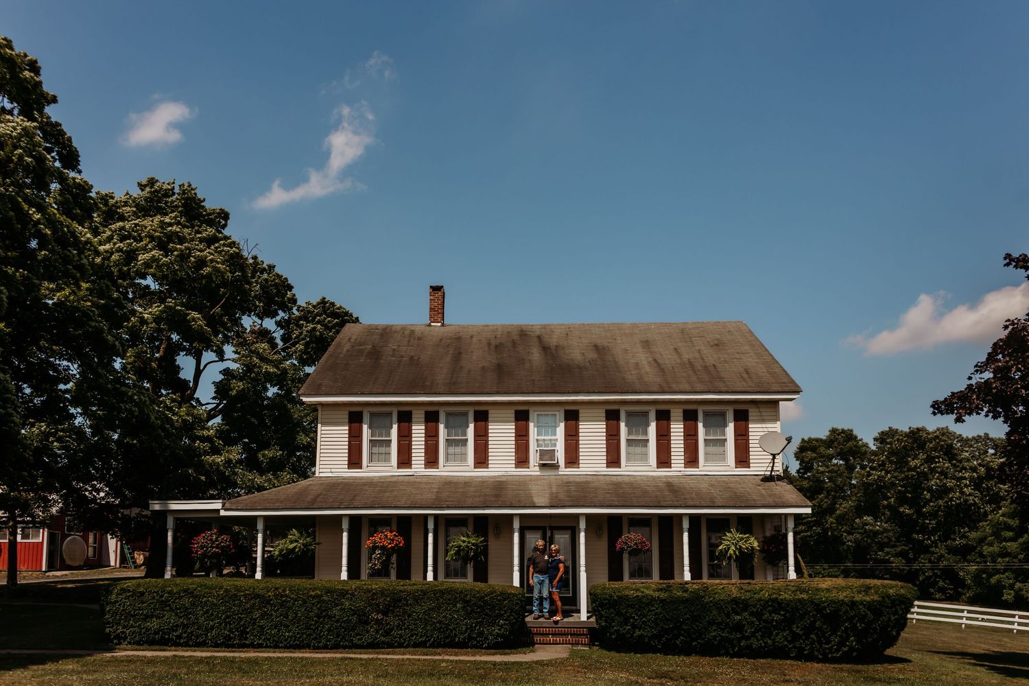 Large white farmhouse with wraparound porch under blue summer sky.
