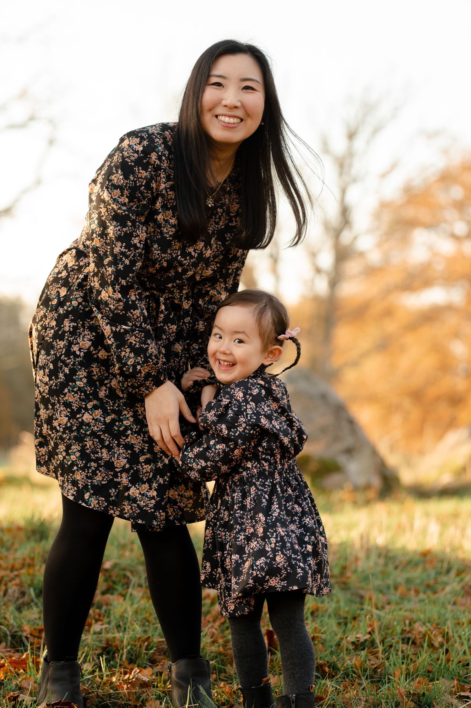 Two people wearing matching floral dresses pose together in an autumn field at sunset.
